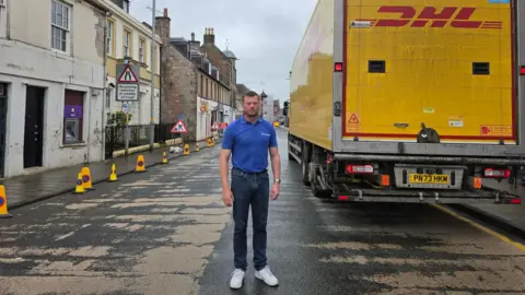 Emma Gilchrist A man in a blue T-shirt and jeans stands in the middle of Sanquhar's main street with a lorry beside him and police traffic cones up the other side of the street.