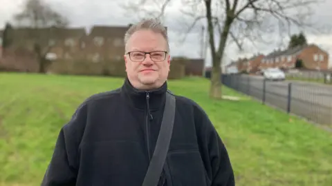 Rick Burgess, who is wearing a dark blue fleece with a bag slung over his shoulder. He is standing in a park on a housing estate in Oldham. 
