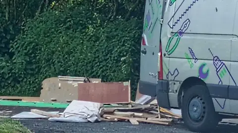 The corner of Boiler Road and Castle Island Way in Ashington, next to the woods at Wansbeck Riverside Park. A van with a livery of cartoon tools - a drill, a pair of pliers, a saw, etc - has its rear doors open. Rubbish, predominantly made up of broken wood, is sprawled across the ground behind it.