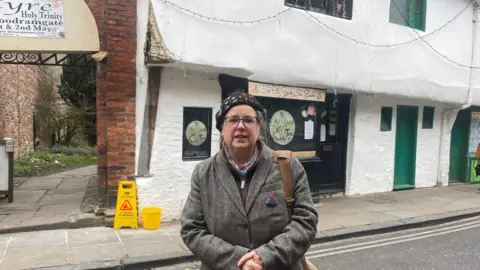 A woman standing on a narrow street in front of a historic-looking building. The building has white plaster walls with a slightly uneven, old-style appearance, and several green-painted doors and windows.