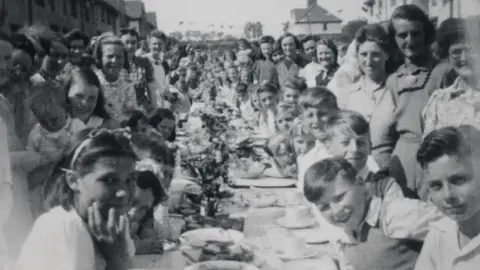 A black and white photo of a street party. There is a long table lined with a tartan cloth. There are white plates and teacups all over the table and in the centre there are flowers going down the line of the table. Lots of children are sat at it, leaning to the camera, surrounded by women holding smaller children.