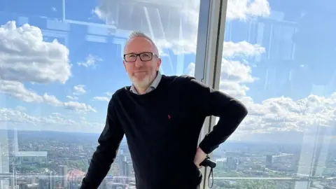 Nick Brooks A man in a black jumper stood in front of a large glass window.