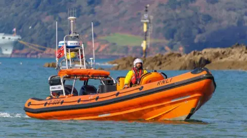 RNLI Will Roberts is at the helm of an RNLI inshore lifeboat, a branded orange rigid inflatable boat. The sea is calm and the rocky shoreline can be seen behind him as well as a part of a large military boat which is moored up. He is wearing RNLI uniform, including a lifebelt and a white helmet.