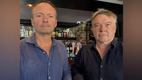 Adrian Rothera and Rory Fry-Stone, co-owners of Pilgrim brewery which runs the Cage Wine Bar in Reigate. Adrian, on the left, is wearing a blue shirt and has greying hair. Rory, who also has greying hair, is wearing a navy shirt.