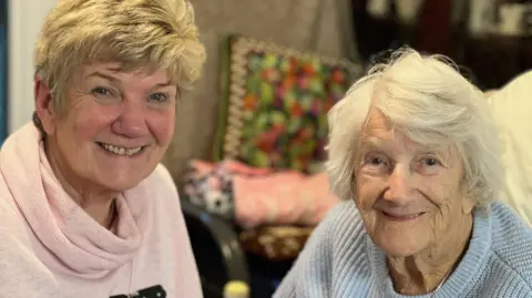 Jane has medium-length grey-blonde hair and is wearing a pale pink jumper. Her mum Betty has grey hair and is wearing a pale blue jumper. Both women are smiling for the camera.