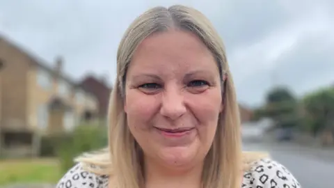 Nicola is smiling, she has brown eyes and freckles dappled across the right side of her cheek. She has a long blonde bob and is standing in front of a road which has been blurred.