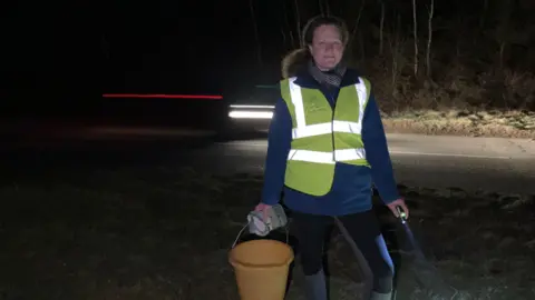 A woman in her 30s with a ginger frizzy ponytail stands on a road side at night, holding a yellow bucket and a torch. She is wearing a hi-vis tabard, a blue jacket, and black leggings. A car with its headlights on can be seen whizzing behind her.