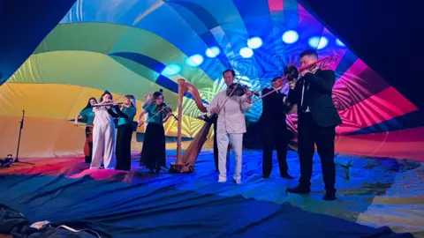 Musicians playing instruments inside a rainbow coloured hot air balloon as it opens up, with lights shining inside it. There are around eight musicians playing violins, a trombone, cello, harp, and clarinet.
The men are wearing white or black smart suits and three of the women are wearing black skirts and turquoise long sleeved silk-style tops and one is in a long sleeved whited top and a long white skirt. They are standing on the rainbow material of the balloon as it surrounds them. 