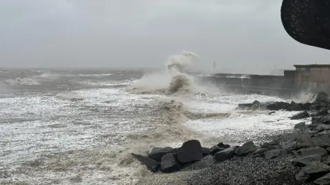 BBC Weather Watchers The seafront in Watchet, Somerset. Big waves can be seen crashing into the sea wall. The sky is very grey, cloudy and bleak. 