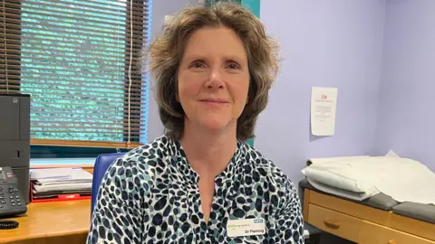 A woman sits in a GP treatment room, behind her is a window and a telephone, to the right of her is a treatment bed. She is smiling, has shoulder length light brown hair and is wearing a turquoise and black patterned blouse with an NHS name tag that reads 'Dr Fleming'.