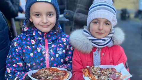 Two young smiling girls with pizza. The youngster on the left is wearing a blue woolly hat and dark jacket with blue, pink and white petals on its design. Her friend has a white woolly hat and a pink coat with white fur-style collar.