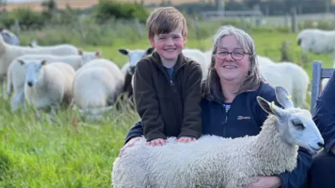 Sally Crowe kneels in a field with her hand around her young son William. They are both wearing fleeces and there is a small sheep standing in front of them.