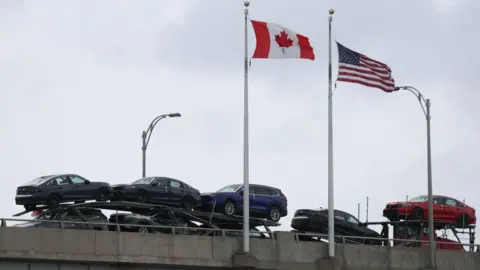 The Canada and US flags flap in the wind on flag polls as a truck stacked with cars for sale drives by