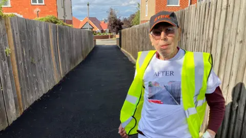 James Grant/ BBC Chris Antoniou in a safety vest and a white T-shirt stands proudly on a freshly paved path