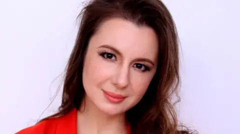 Dr Anna Kruglova A woman with long brown hair smiles at the camera. She is wearing a red top and the background is off-white.