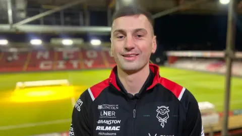 Footballer Paudie O'Connor stands in the Lincoln City FC stadium. He is smiling and short brown hair. He is wearing a black club tracksuit top with red trim which covered in sponsor logos and the football club badge. The green football pitch is visible in the background alongside the red and white-coloured seats.