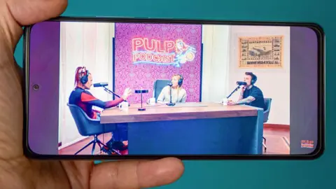 Getty Images A hand holds a phone in landscape playing the stream of the Pulp podcast featuring an interview with Giorgia Meloni. The hosts are two young men, one of whom has tattoo sleeves and the other has red dreadlocks. Meloni is wearing a jumper. The three of them are sat around a table with a microphone and headphones. 