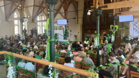 Notting Hill Methodist Church, Lancaster Road. Photo shows people sat in wooden pews and the columns are decorated in green and white flowers. 