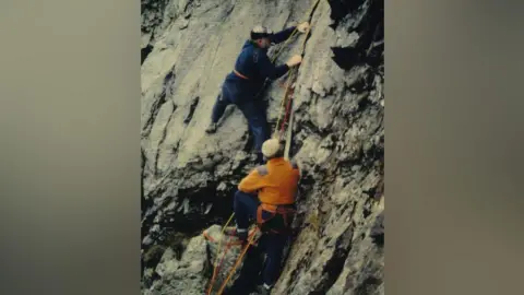 Bill Birkett Bill Peascod and Don Whillans climb Eagle Front above Buttermere. Two men stand on a rocky crag. Both are wearing jeans and flat caps, one man wears an orange jumper and the other wears a navy jumper.