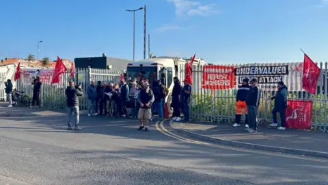 Striking workers picket outside the front of a depot. They are blocking an entrance between silver fences. Some are holding red Unite flags. A lorry can be seen parked up in the entrance.
