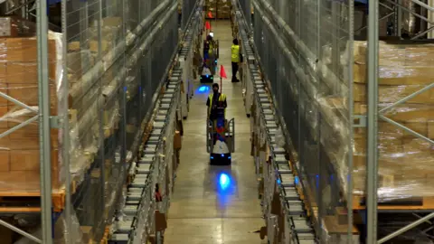 DP World A long corridor between enormous shelves covered with boxes. In the corridor, white robots move around alongside staff in hi-vis jackets.