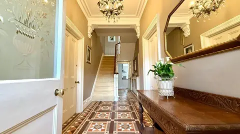 A hallway into the crisis house - before renovations started - featuring Victorian-patterned brown floor tiles, a frosted glass door, stained wooden furniture, a white staircase and multiple chandeliers.