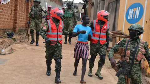 Anadolu via Getty Images A man in blue jersey and stripped shorts is accosted by security forces in military uniform in Kampala