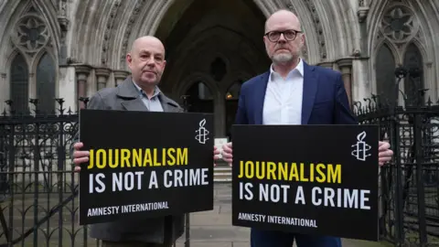 Barry McCaffrey and Trevor Birney outside the Royal Courts of Justice, London, in October. They are holding black signs with yellow and white writing that say 'Journalism is not a crime'. There are black iron gates behind them.