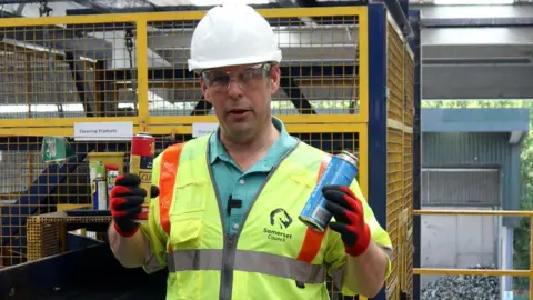 A man pictured in a high vis sleeveless jacket, safety goggles and a white hard helmet holding up two gas canisters. One is yellow and red, the other is blue. He is handling them in red gloves. 