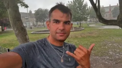 Facebook Tancredo Bankhardt - a man with short dark hair, standing outside near a park taking a selfie. He is looking directly at the camera and is wearing a grey T-shirt with a necklace. His left hand is in the air, with his thumb and little finger outstretched.