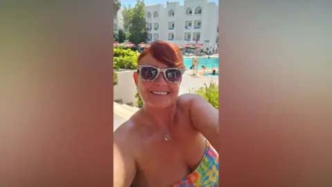 Martine Murray A woman is taking a selfie at a holiday resort. In the background is a swimming pool. She has ginger hair and is wearing a bikini. 