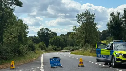Moy road. Two yellow police cones are on the road with a blue 'Police, Accident' sign. To the right is a PSNI are with the door open. Road is surrounded by green trees.