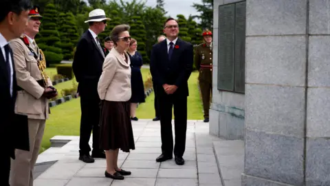 PA Media Princess Anne pictured at a memorial cemetery in Busan. She is pictured wearing sunglasses, a cream jacket and a brown dress while looking at a memorial full of the names of deceased soldiers.