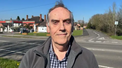 A man stood next to a road in a grey jacket and a checked shirt, smiling at the camera.