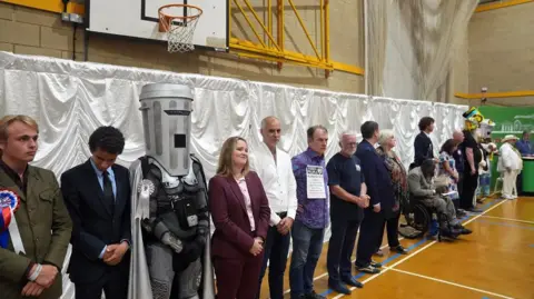PA Candidates listen to the declaration for the Clacton by-election count at Clacton Leisure Centre in Essex. The by-election was triggered by local MP and Reform UK leader Nigel Farage quitting in protest at the scrutiny of his finances from the press and the parliamentary authorities. Picture date: Friday August 14, 2026. PA Photo. Photo credit should read: Joe Giddens/PA Wire