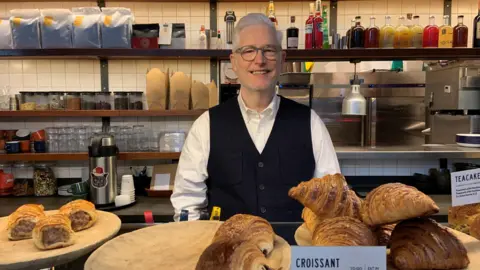 A man has white hair in a quiff and is wearing glasses, a white shirt and black waistcoat. He is standing behind a counter with several croissants, in the background are shelves with coffee bags, a coffee machine, glasses and bottles
