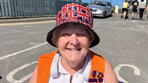 A woman smiles at the camera while enjoying an Orange Order parade on a sunny day in County Donegal. She is wearing a hat decorated with small union flags, a white sleeveless top and an orange collarette