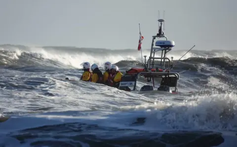 Helen Cowan An orange lifeboat disappears among large waves. Just visible are the top halves of four crew members wearing yellow dry suits and white helmets.
