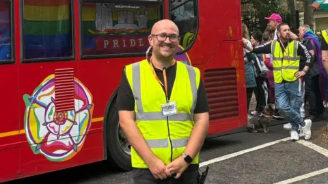 A person in a high‑visibility yellow vest stands in front of a red bus decorated with colorful graphics and the word "PRIDE".