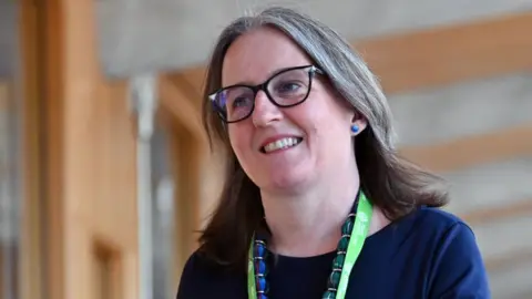 Getty Images Maree Todd walking down a corridor in the Scottish Parliament. She has shoulder-length hair and glasses, and is wearing a blue top, a blue and green necklace and a lanyard with a green strap.