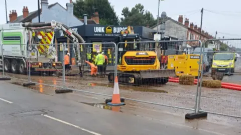 A metal fence and orange cones surround a wet road and workers in hard hats and yellow and orange jackets survey the scene