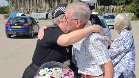 Wiltshire Police 74 year old David Painter with grey hair, glasses and striped short-sleeved shirt hugging PC Vicky Howick in police uniform and holding a bouquet of pink and white flowers. 
