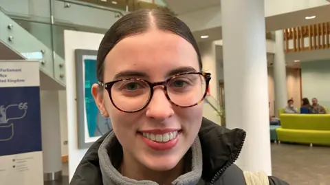 Student Jessica Skelly is wearing a grey fleece and a black coat. 
She is smiling and is wearing glasses with her brown hair parted in the middle. 