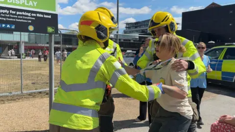 A blonde-haired woman with sunglasses on, a white top and black trousers is being held up by three people dressed in yellow high-vis and helmets. She has a shocked looked on her face. A few people are in the background, along with a police vehicle and a sign that says student's union.