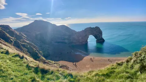 The rock arch of Durdle Door on the Dorset coast.  The cliff juts out into the water and a bright blue sea. The sun is low in the blue sky. In the foreground you can see clifftop grass and on the beach below are several people. 