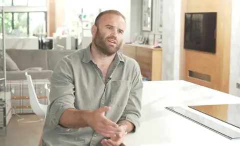 Former Welsh international rugby player Jamie Roberts sits in his kitchen talking to the reporter. He is wearing a grey shirt.