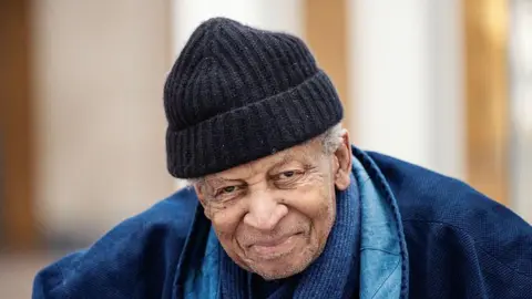 Abdullah Ibrahim in a black beanie hat and blue jacket smiles as he looks at the camera.