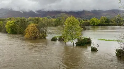 Paul Rathbone A flooded area of land near Keswick. The trees are deep into the brown floodwater. Thick grey cloud looms over the fells in the background. 