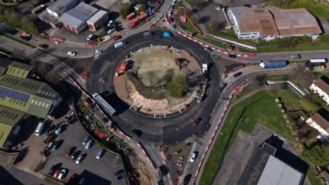 Drone image taken above a large roundabout where roadworks are taking place. There are four exits. Vehicles can be seen driving around the roundabout in lanes denoted by orange cones. The middle of the roundabout has been dug up and there are construction vehicles on it. An industrial and business area surrounds the roundabout.