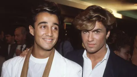 Getty Images Archive photo of Andrew Ridgley and George Michael of Wham! Andrew is where a white top, George is where a shite short and dark jacket and is looking at the camera.